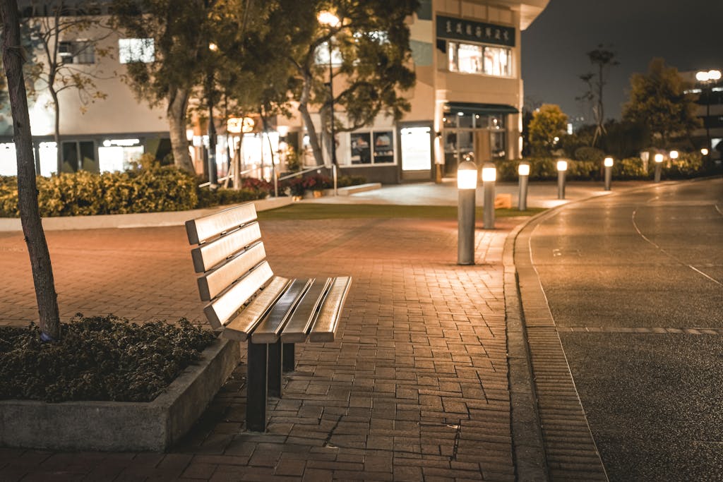A serene city street scene featuring an empty bench at night, illuminated by streetlights.
